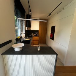 GLAZERIS Banakiewicz Adrian - Nowoczesna kuchnia z czarnym blatem i białymi frontami szafek, oświetlona designerskimi lampami. Widoczne okno z żaluzjami i drewniane elementy wykończenia.