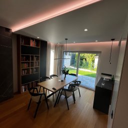 GLAZERIS Banakiewicz Adrian - Nowoczesna jadalnia z drewnianym stołem, krzesłami i regałem na książki. Subtelne oświetlenie LED podkreśla minimalistyczny design wnętrza z widokiem na ogród.