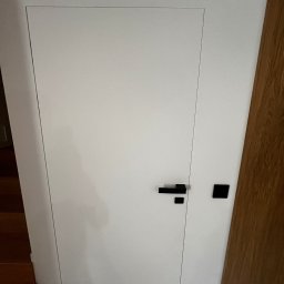 GLAZERIS Banakiewicz Adrian - Białe, minimalistyczne drzwi ukryte z czarną klamką i zamkiem na tle białej ściany i fragmentu drewnianej okładziny. Widoczne schody w tle.