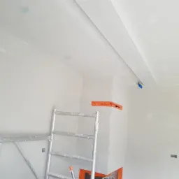 Wnętrze w trakcie prac wykończeniowych: sufit z listwami dekoracyjnymi, ściany po gładziowaniu, widoczne rusztowanie i narzędzia.