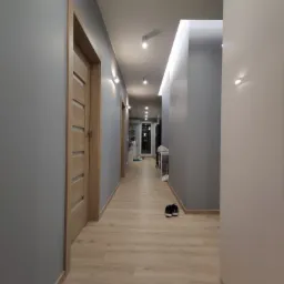 Korytarz po remoncie z drewnopodobną podłogą, szarymi ścianami i drewnianymi drzwiami, oświetlony punktowymi lampami i ledowym pasem na suficie.