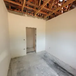 Pokój po nałożeniu gładzi gipsowej, widoczna drewniana konstrukcja dachu, przygotowany otwór na drzwi, instalacja elektryczna wypuszczona ze ściany.