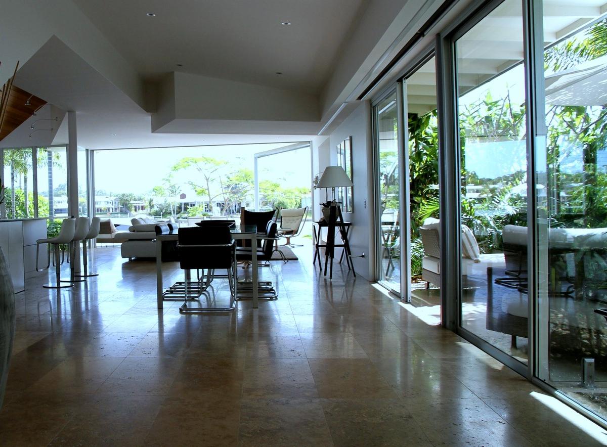 Przestronne, nowoczesne wnętrze domu z dużymi przesuwnymi oknami wychodzącymi na zielony ogród i wodę, polerowana kamienna podłoga odbija światło, minimalistyczne meble.
