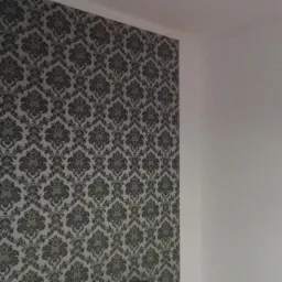 Ściana pokryta tapetą w ornamenty kwiatowe w odcieniach szarości, sąsiadująca z białą ścianą i oknem z żaluzjami plisowanymi.