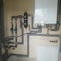 Nowoczesny piec gazowy Viessmann z rozbudowaną instalacją hydrauliczną i grzewczą, rury zaizolowane szarą pianką, widoczne zawory, pompy obiegowe i manometry, ściany w surowym stanie.