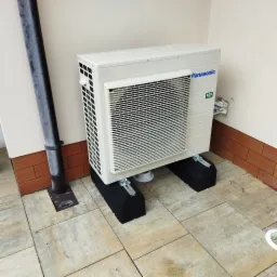 Zewnętrzna jednostka klimatyzacji Panasonic R32 na czarnych podstawach, zamontowana przy ścianie z brązową cegłą i rurą spustową.