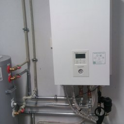 Budwar Skłodowski Robert - Biała pompa ciepła Aquarea Smart Cloud zamontowana na ścianie, widoczne rury doprowadzające wodę i elementy instalacji hydraulicznej w pomieszczeniu technicznym.