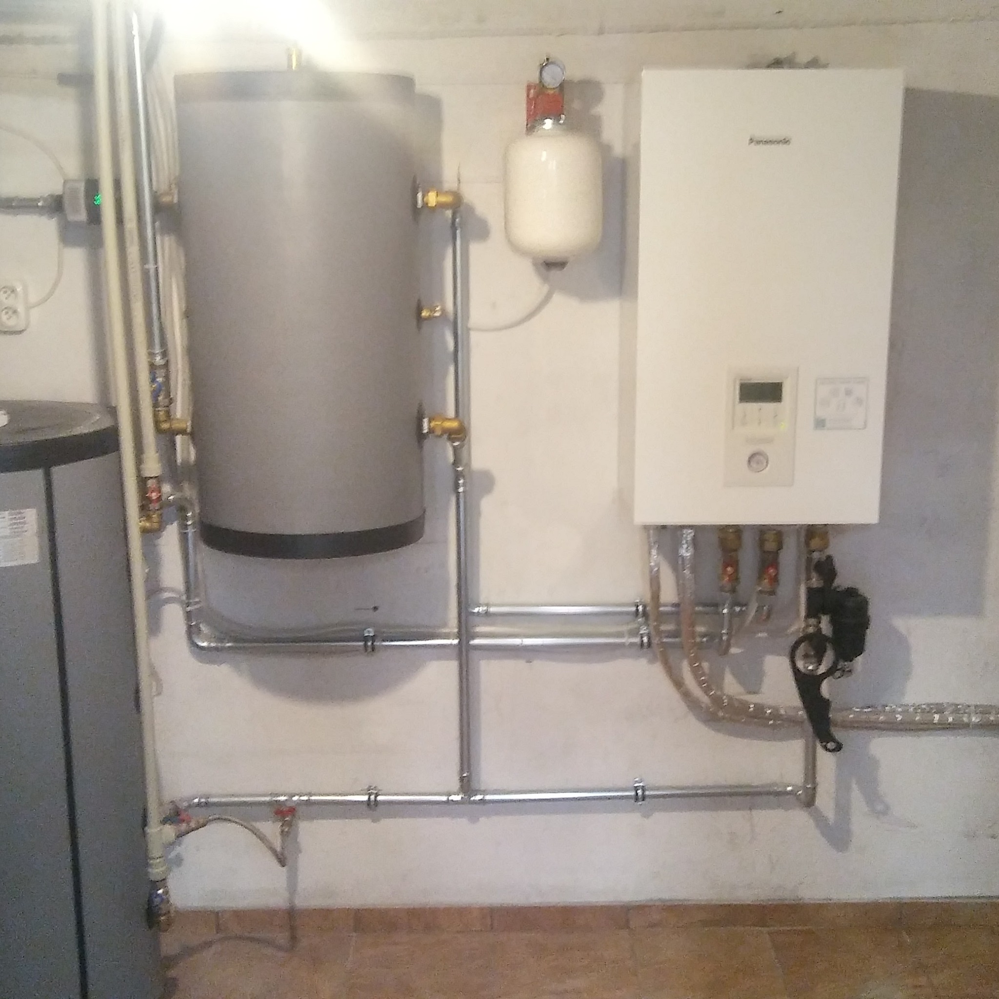 Instalacja grzewcza z szarym zbiornikiem akumulacyjnym, białym zasobnikiem ciśnieniowym i kotłem gazowym marki Panasonic na ścianie w piwnicy.