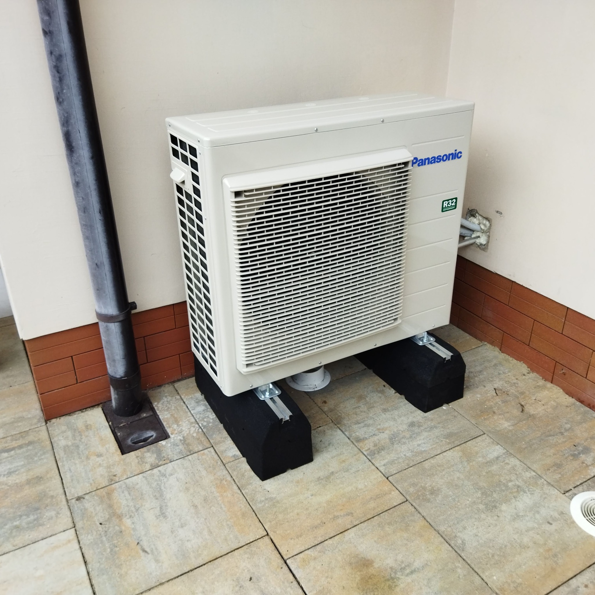 Zewnętrzna jednostka klimatyzacji Panasonic R32 na czarnych podstawach, zamontowana przy ścianie z brązową cegłą i rurą spustową.