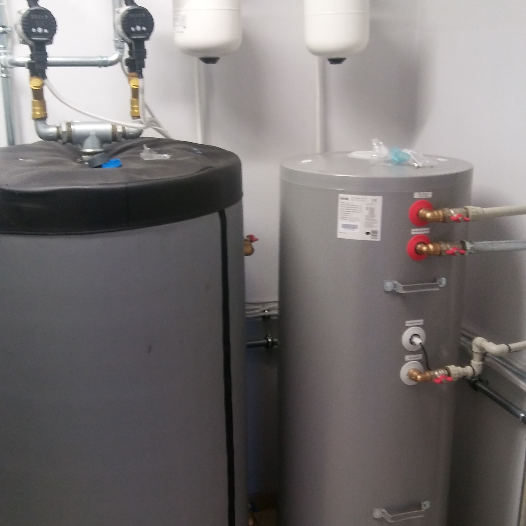 Dwa szare zbiorniki, jeden z czarną izolacją i podłączonymi rurami z zaworami, drugi z etykietą i czerwonymi uszczelkami wokół przyłączy rur.