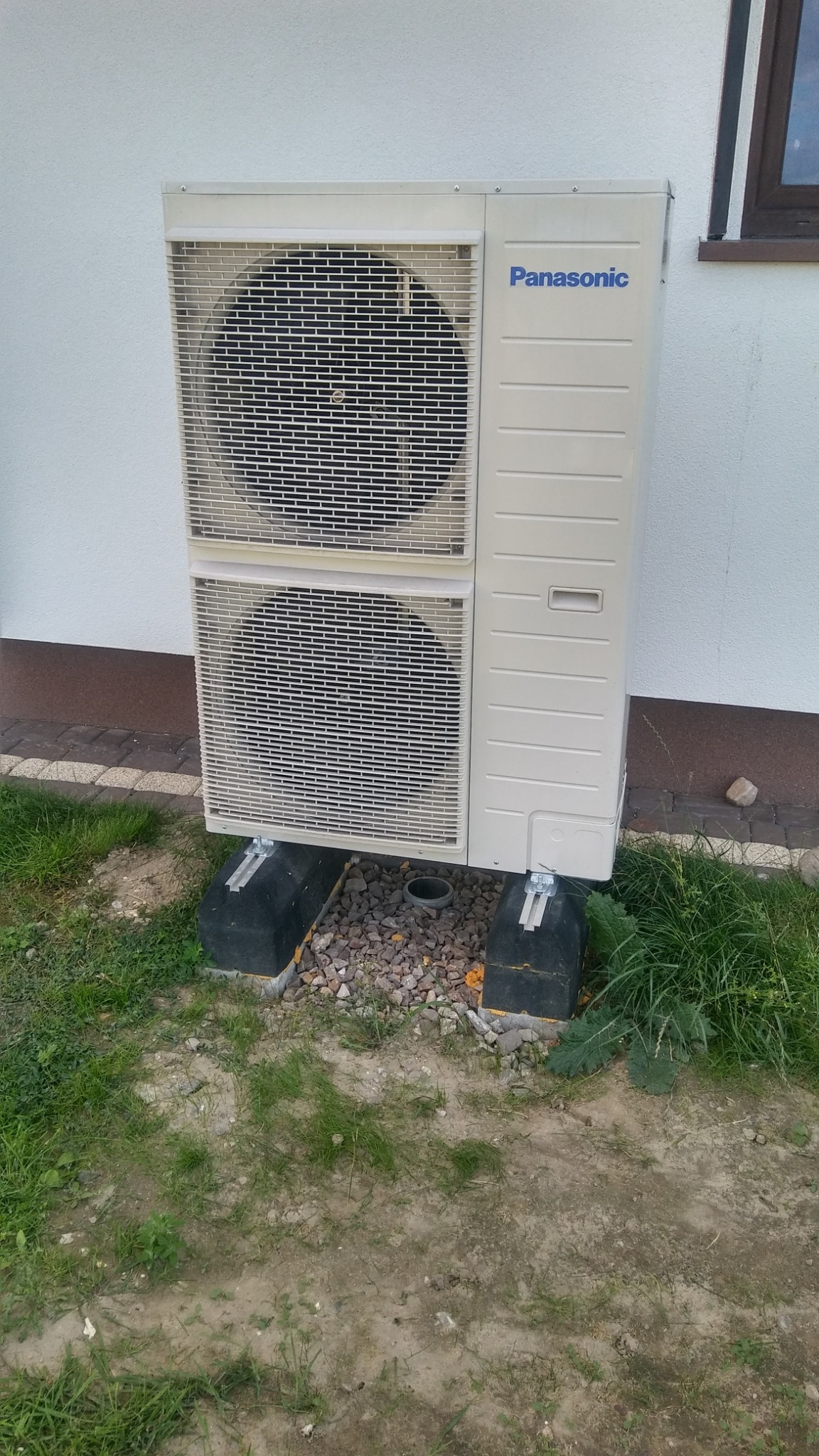 Zewnętrzna jednostka klimatyzatora Panasonic zamontowana na betonowych podstawach z podsypką żwirową przy ścianie budynku z widocznym oknem na piętrze.