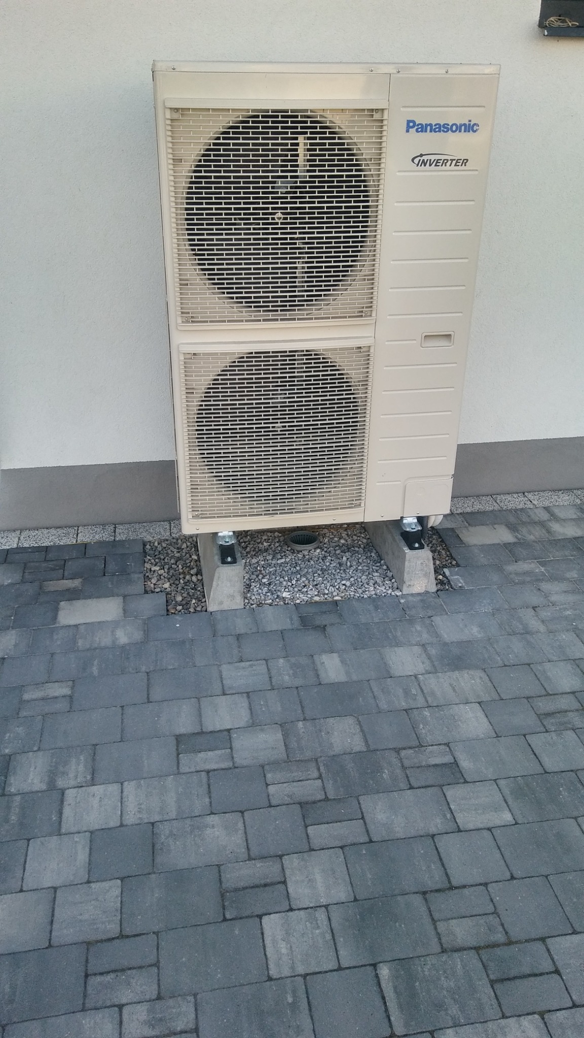 Zewnętrzna jednostka klimatyzatora Panasonic Inverter zamontowana na betonowych podstawach z tłuczniem, widoczna na tle ściany budynku i szarej kostki brukowej.