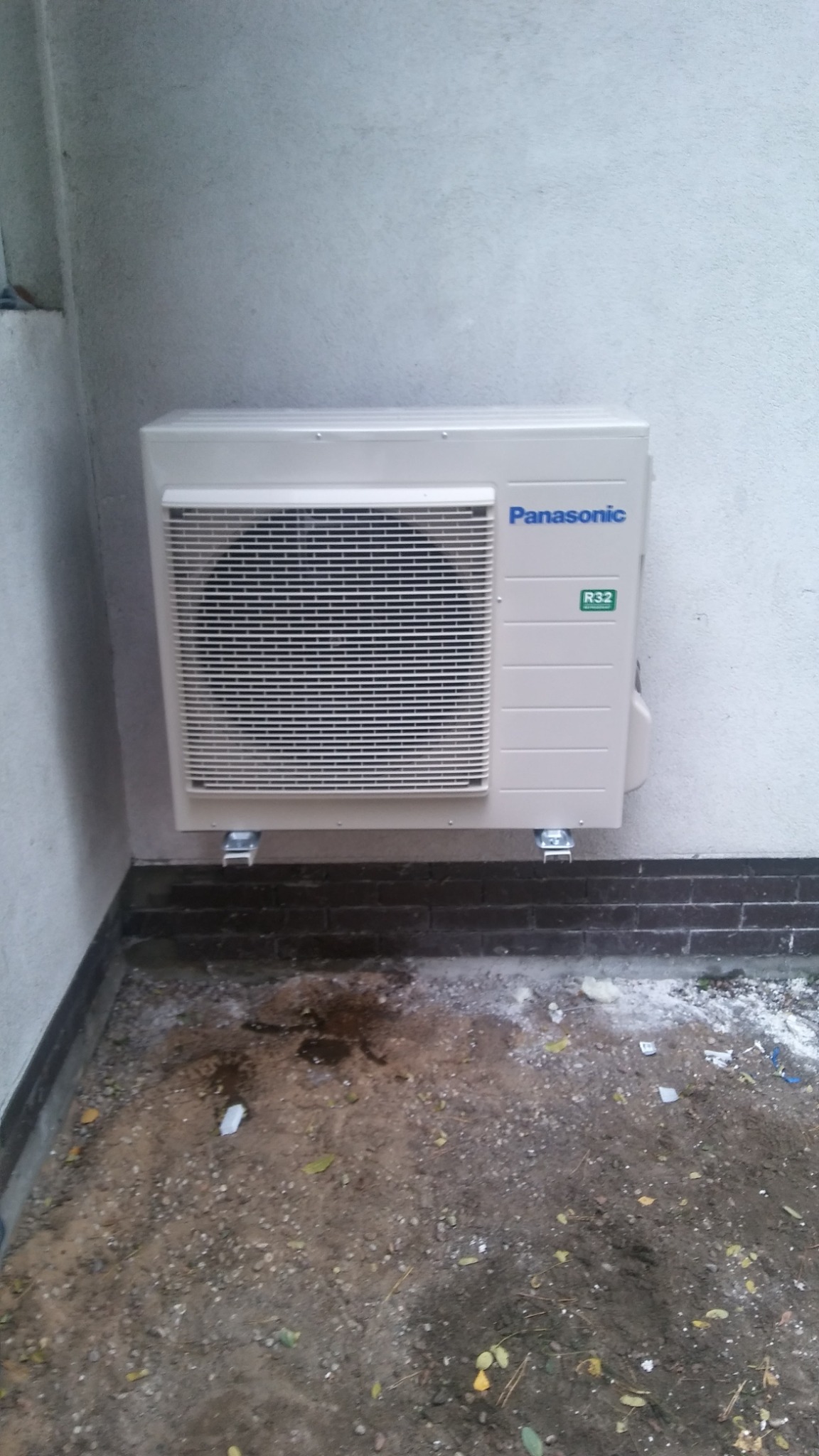 Zewnętrzna jednostka klimatyzacji Panasonic zamontowana na ścianie budynku, widoczne ślady montażu na podłożu.