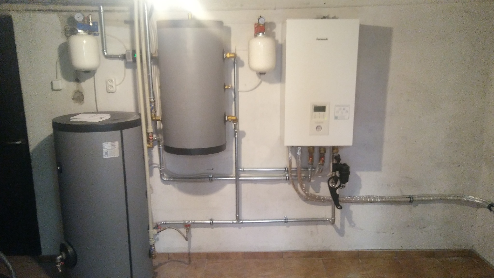 Kotłownia z zainstalowanym zbiornikiem akumulacyjnym, pompą ciepła Panasonic i osprzętem hydraulicznym, widoczne rury i manometry.