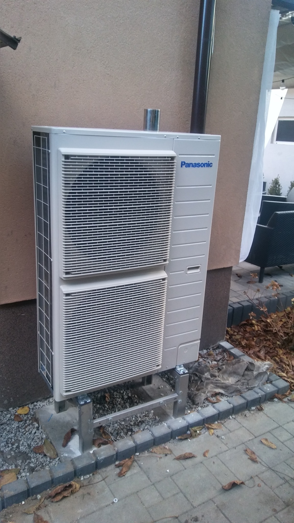Zewnętrzna jednostka klimatyzacji Panasonic zamontowana na metalowej konstrukcji przy ścianie budynku, otoczona kostką brukową i opadłymi liśćmi.