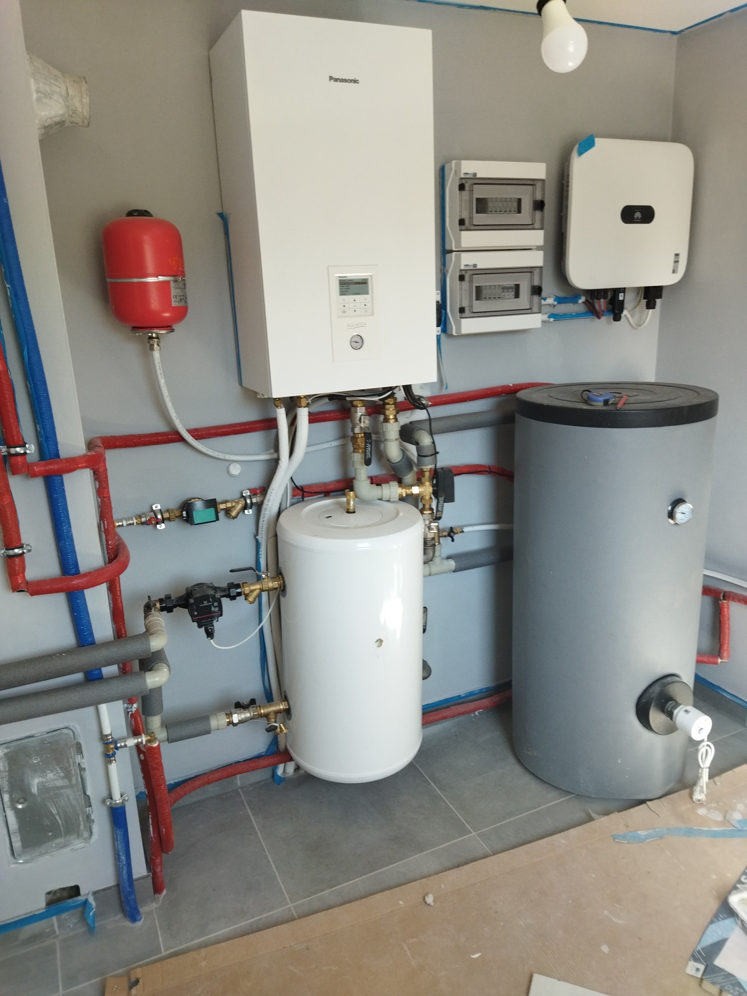 Instalacja grzewcza w kotłowni: biały zbiornik akumulacyjny Panasonic, szary zasobnik ciepłej wody, czerwony zbiornik przeponowy, liczniki i inwerter na szarej ścianie, widoczne rury miedziane...