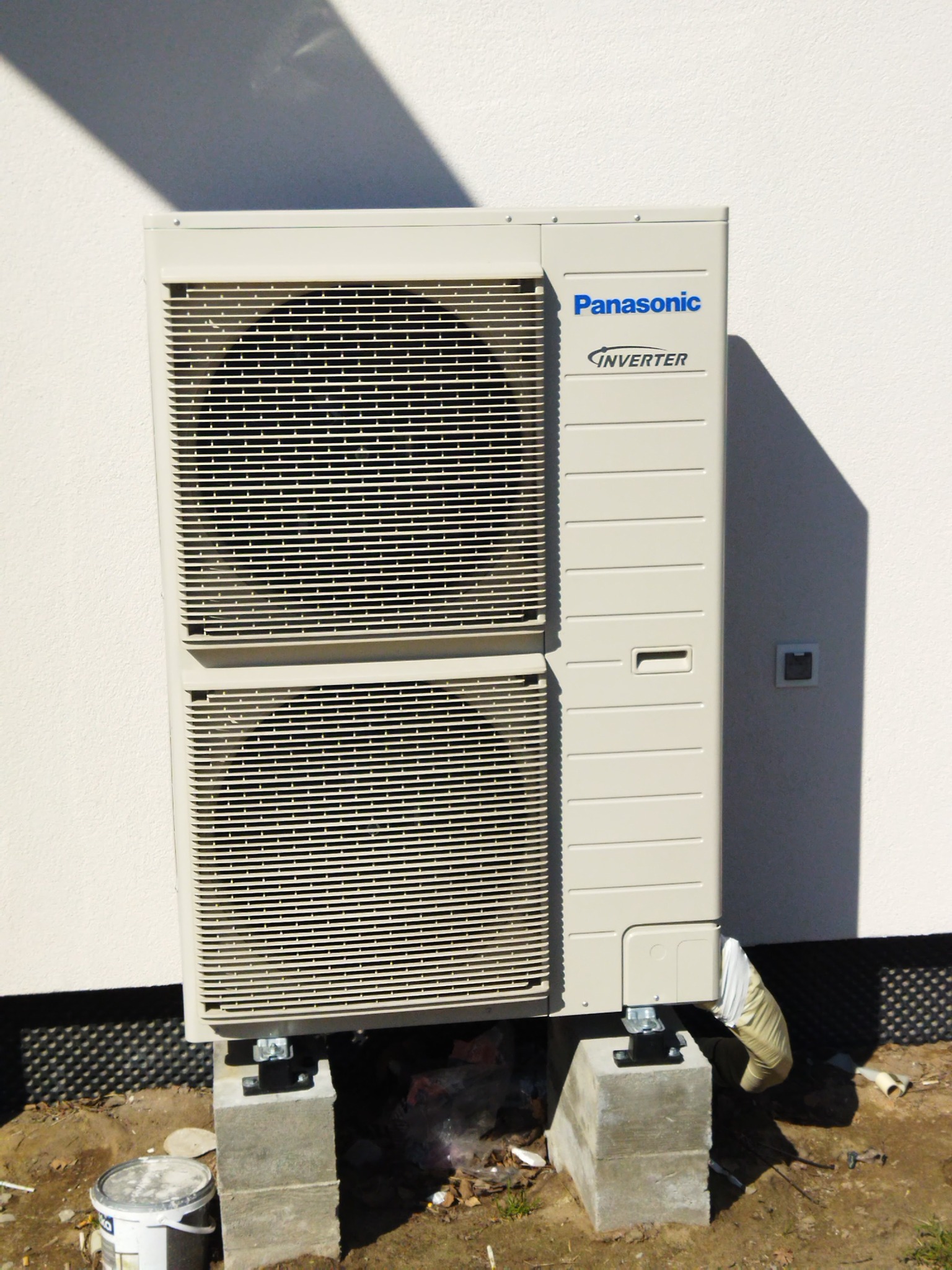 Zewnętrzna jednostka klimatyzacji Panasonic Inverter zamontowana na betonowych bloczkach, z widoczną izolacją rury odprowadzającej skropliny, na tle jasnej ściany budynku z widocznym cieniem.