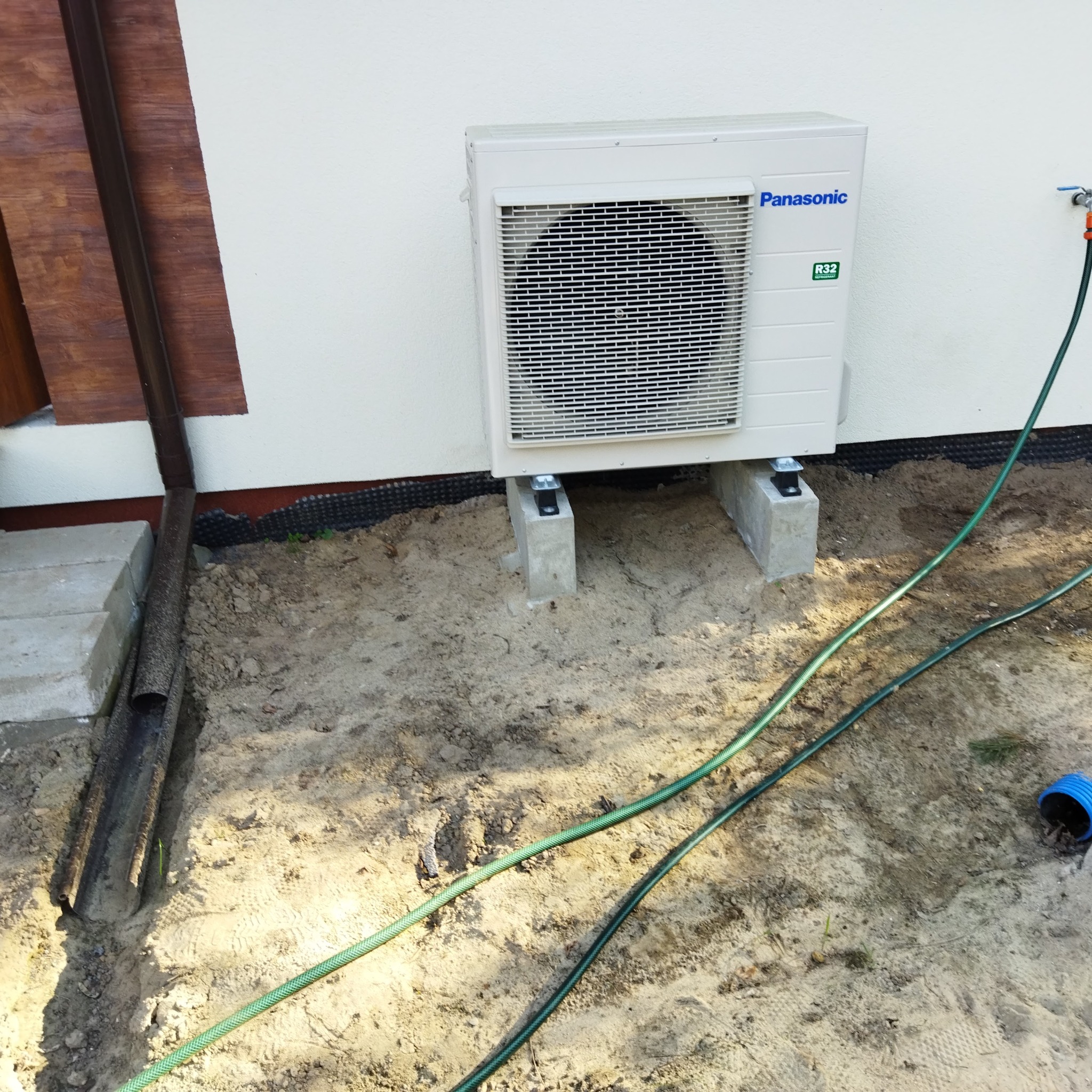 Zewnętrzna jednostka klimatyzacji Panasonic R32 zamontowana na betonowych podstawach, obok rury spustowej, na piaszczystym podłożu z widocznymi wężami i przyłączem wody.