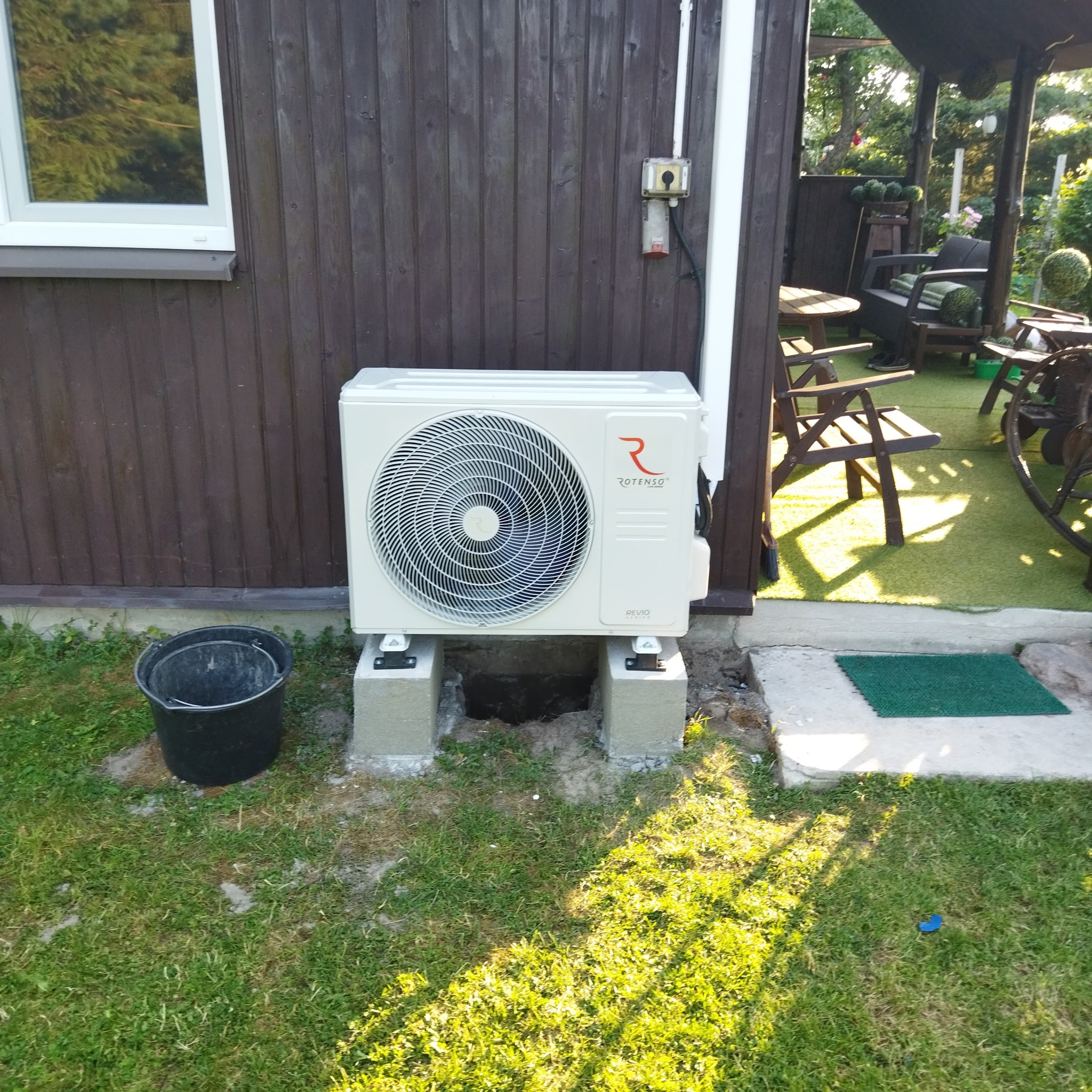 Biała jednostka zewnętrzna klimatyzacji Rotenso Revio zamontowana na betonowych postumentach przy drewnianej ścianie budynku, obok czarne wiadro i fragment tarasu z meblami ogrodowymi.