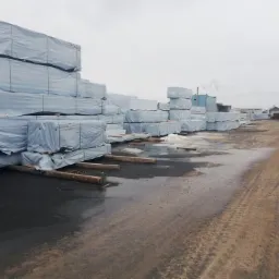 Sztaple tarcicy budowlanej owiniętej folią na terenie składu, widoczna osoba w kurtce i jeansach, mokra nawierzchnia placu.