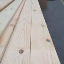 Stos jasnej tarcicy sosnowej z widocznymi sękami, ułożonej w magazynie z betonową podłogą i innymi materiałami budowlanymi w tle.