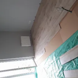 Montaż jasnych paneli podłogowych w pokoju z szarą ścianą i grzejnikiem; zabezpieczenie podłoża folią ochronną; widoczne przewody elektryczne wystające z podłogi.