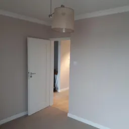 Pomalowane na beżowo ściany pokoju z białymi listwami przypodłogowymi i sufitowymi, białe drzwi wewnętrzne z srebrną klamką, widok na parkiet w przejściu do kolejnego pomieszczenia, wisząca lampa...