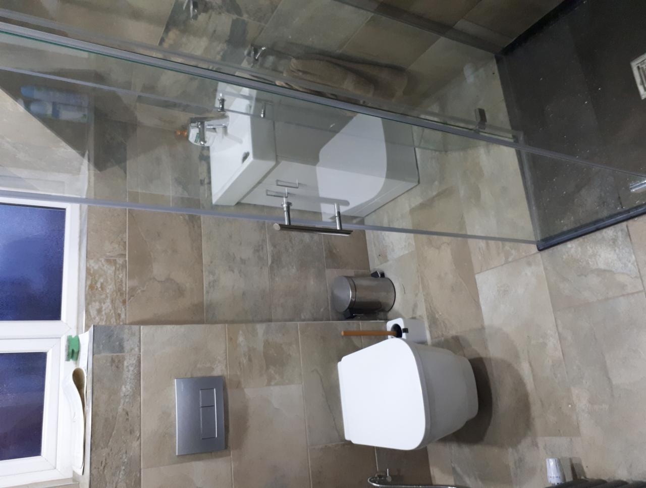 Nowoczesna łazienka z beżowymi płytkami imitującymi kamień, kabiną prysznicową ze szkła hartowanego, wiszącą toaletą i umywalką, oraz chromowanymi akcesoriami.