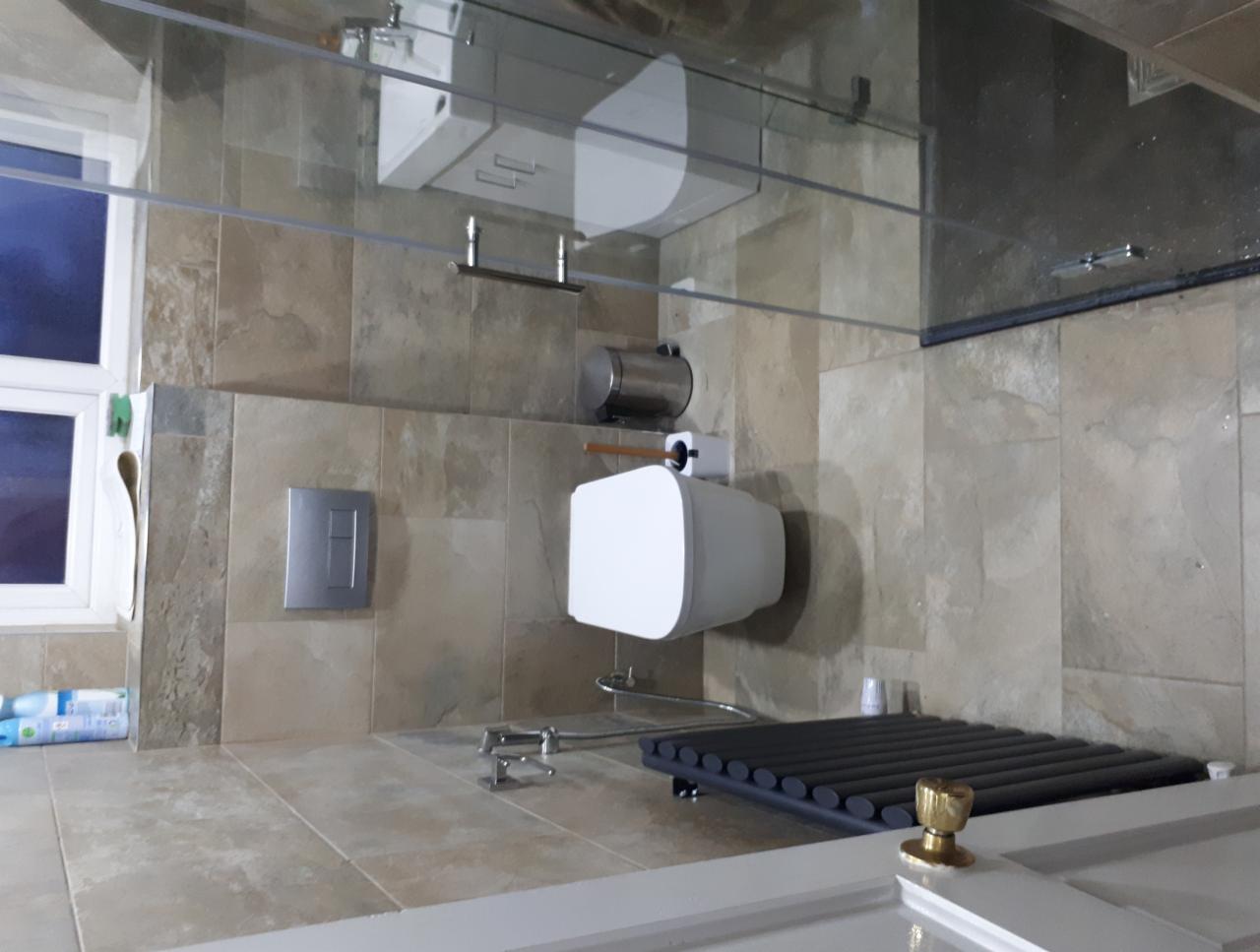 Nowoczesna łazienka z beżowymi płytkami, podwieszaną toaletą i grzejnikiem drabinkowym w kolorze antracytowym, widok zza częściowo otwartych białych drzwi.