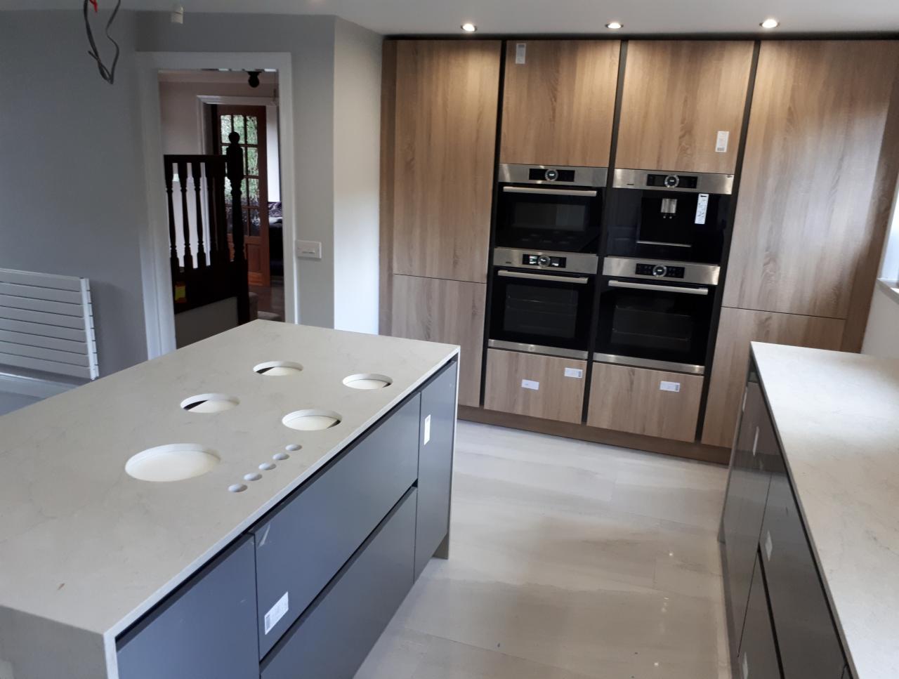 Nowoczesna kuchnia w trakcie montażu: wyspa z jasnym blatem z otworami na zlew i akcesoria, zabudowa kuchenna z drewnopodobnymi frontami i wbudowanymi piekarnikami, jasna podłoga, widok na schody...