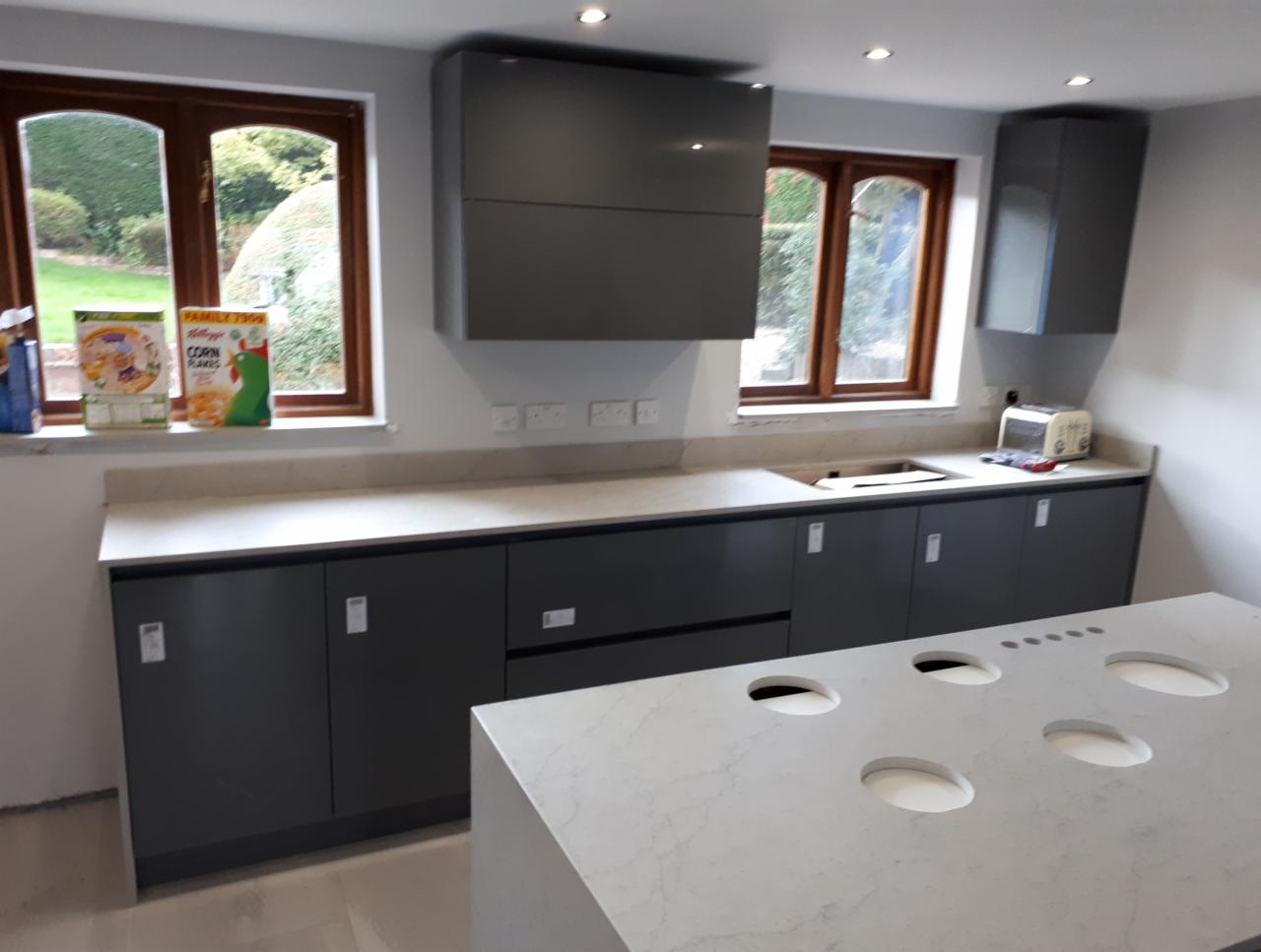 Szara zabudowa kuchenna z wyspą i jasnym blatem, widoczne okna z drewnianą ramą i pudełka płatków śniadaniowych na parapecie.