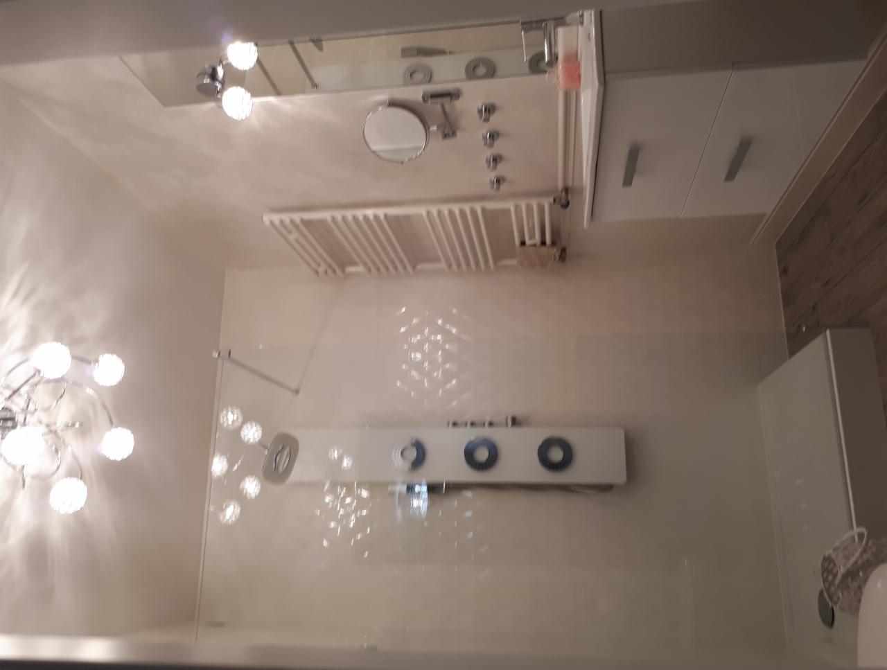 Nowoczesna łazienka z przeszkloną kabiną prysznicową, panelami natryskowymi z okrągłymi dyszami i grzejnikiem drabinkowym, oświetlona dekoracyjnym żyrandolem.