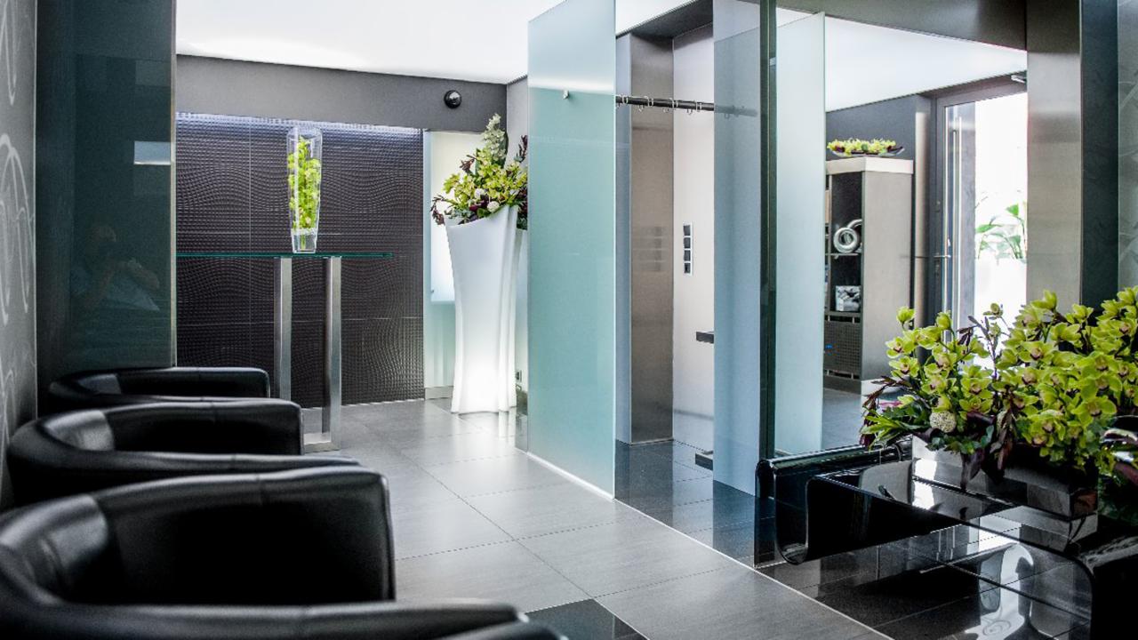 Eleganckie wnętrze holu z czarnymi skórzanymi fotelami, szklanymi ścianami i dekoracjami roślinnymi, prowadzące do windy.