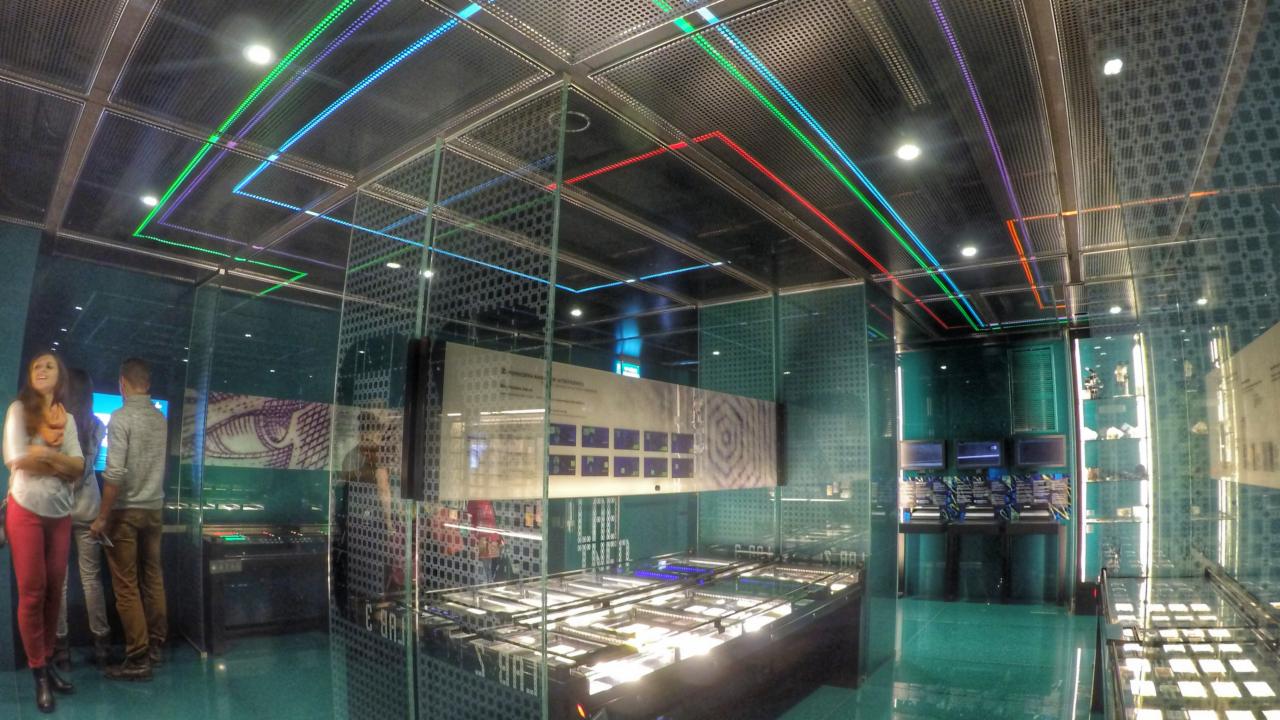Nowoczesne wnętrze muzeum z podwieszanym sufitem z perforowanej blachy i wielokolorowym oświetleniem LED w postaci linii. Na pierwszym planie przeszklone ekspozytory, w tle zwiedzający.
