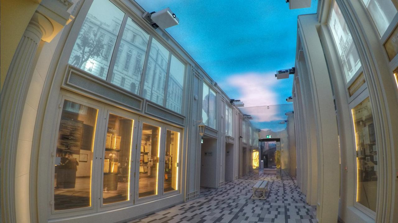 Korytarz w stylu klasycystycznym z kolumnami, oknami z ekspozycjami i podwieszonymi projektorami, sufit imitujący niebo z chmurami.