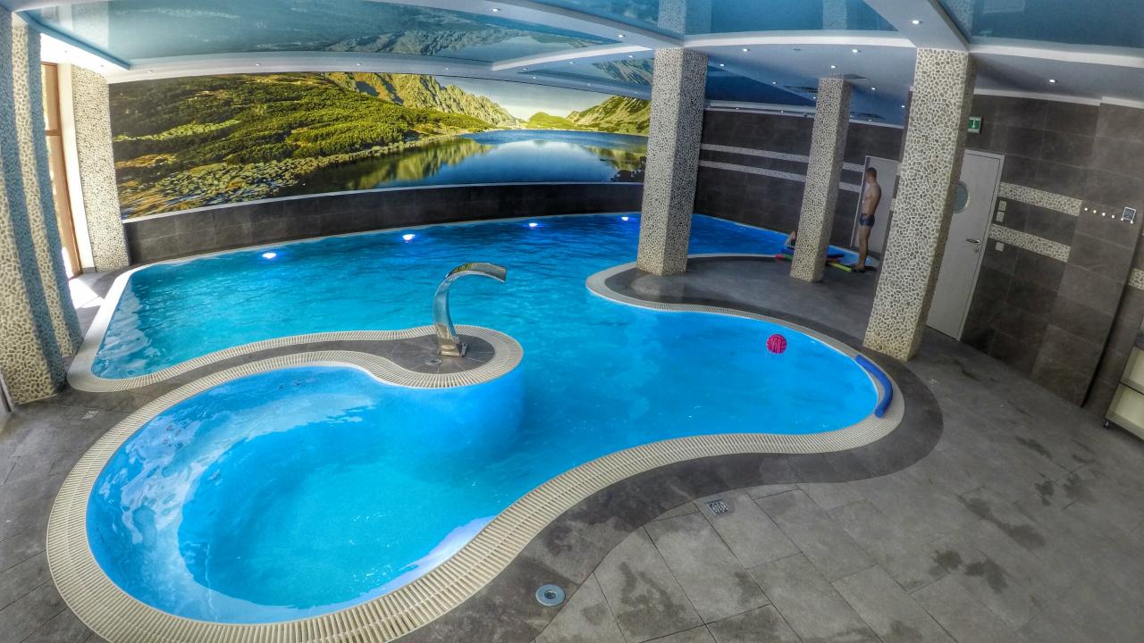 Kryty basen rekreacyjny z dekoracyjną fontanną, mozaikowymi kolumnami i fototapetą górskiego krajobrazu na ścianie.