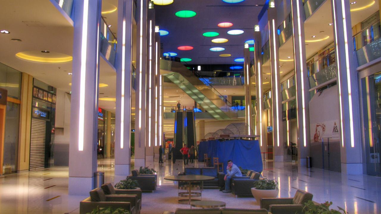 Wnętrze centrum handlowego z wysokimi kolumnami oświetlonymi pionowymi lampami LED, kolorowe okrągłe lampy na suficie, ruchome schody w tle i strefa relaksu z sofami