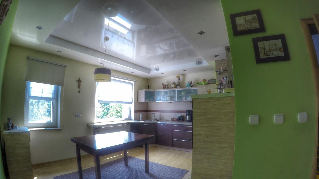 Błyszczący, biały sufit napinany z oknem dachowym w kuchni, widok na stół i zabudowę kuchenną z brązowymi szafkami.