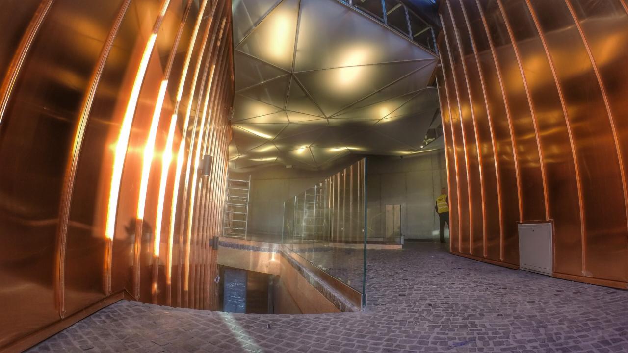 Wnętrze budynku z miedzianymi, żebrowanymi ścianami, geometrycznym sufitem i kamienną podłogą; widoczna drabina i pracownik w odblaskowej kamizelce w tle.