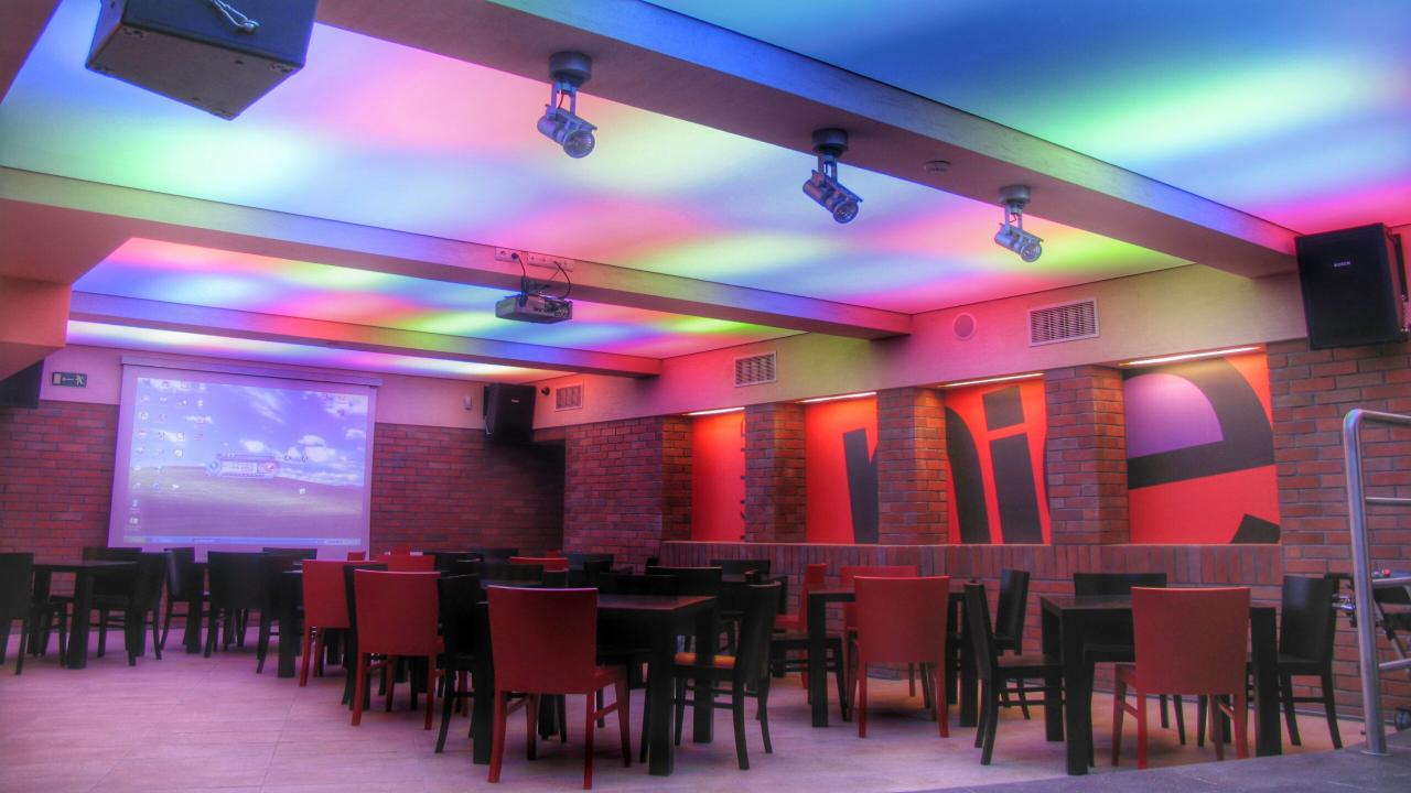 Sala konferencyjna z rzędami krzeseł i stołów, oświetlona kolorowymi reflektorami sufitowymi, z projektorem i ekranem wyświetlającym pulpit Windows XP.