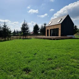 Nowoczesny, parterowy dom z czarnymi ścianami i drewnianą elewacją, z dużymi oknami i drewnianym tarasem, otoczony zielonym trawnikiem i młodymi drzewami iglastymi w słoneczny dzień.