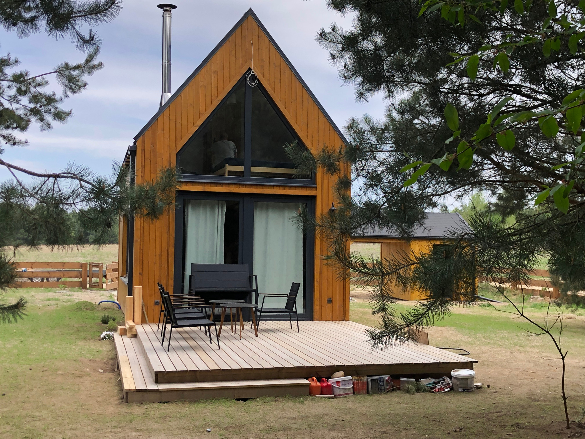 Nowoczesny dom szkieletowy z drewnianą elewacją, dużym trójkątnym oknem na poddaszu, stalowym kominem i przylegającym tarasem z czarnymi meblami ogrodowymi, widok zza drzew z narzędziami...