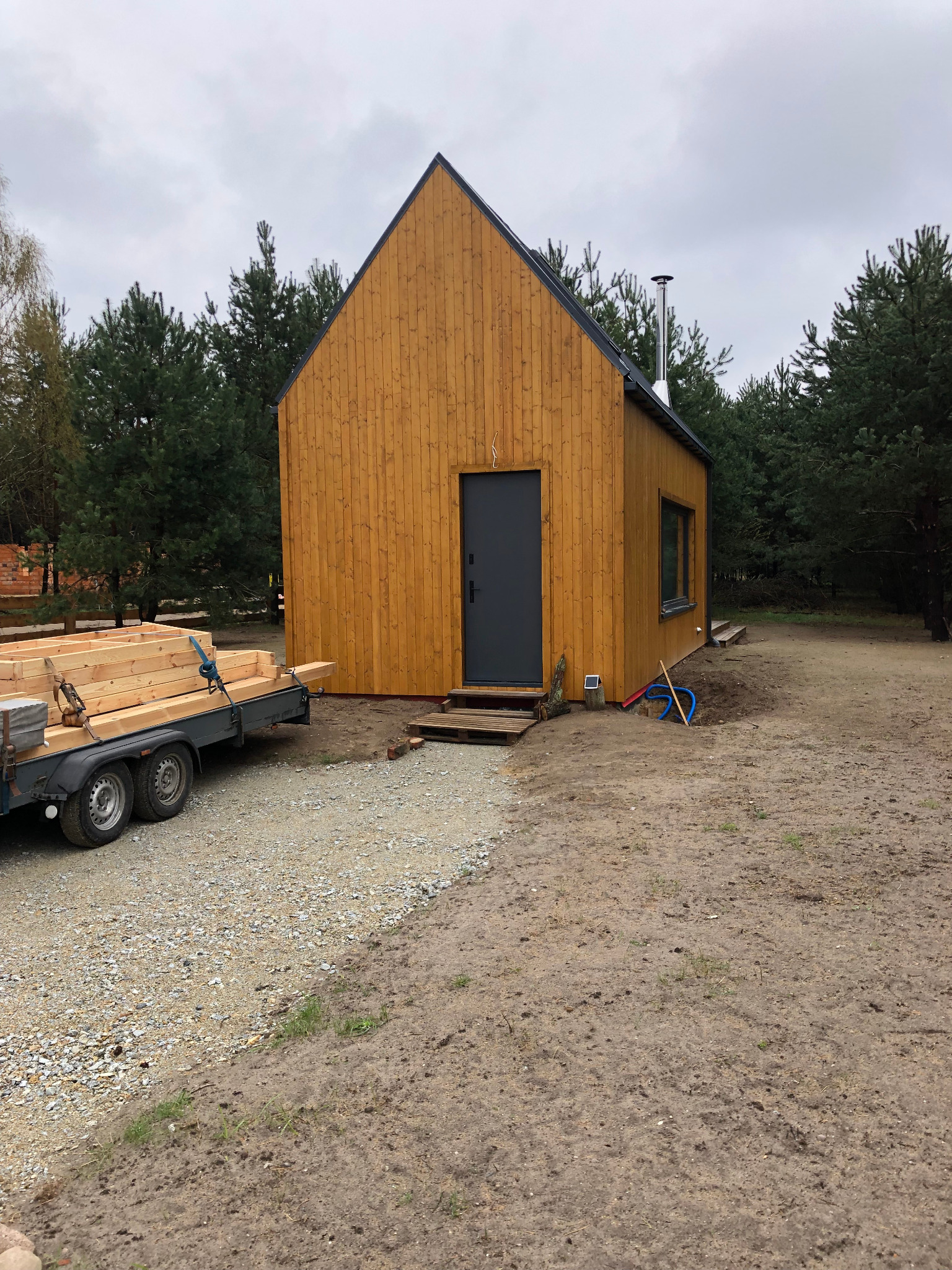 Mały, nowoczesny dom o prostej bryle z drewnianą elewacją, ciemnymi oknami i drzwiami, z kominem, w trakcie budowy, z przyczepą pełną drewnianych belek obok, na działce z drzewami w tle.