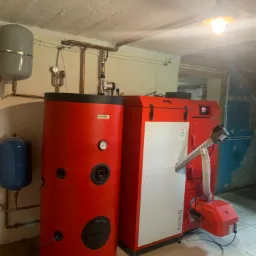 Czerwony piec na pellet DasPell GL i zbiornik buforowy w kotłowni z surowym betonowym sufitem i widocznymi instalacjami hydraulicznymi.