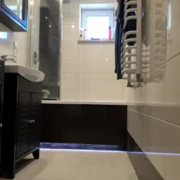 Wykończona łazienka z kabiną prysznicową, szafką pod umywalką z ciemnego drewna, grzejnikiem drabinkowym na ścianie z jasnych płytek i podświetleniem LED pod zabudową wanny.