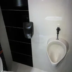 Biały pisuar z chromowaną armaturą na tle białej i czarnej ściany z płytek, obok suszarka do rąk.