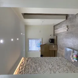 Wnętrze w trakcie wykańczania: ściana z dekoracyjnej cegły, podłoga wyłożona płytkami imitującymi drewno, widoczne materiały budowlane i narzędzia, okno z widokiem na zewnątrz.