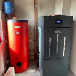 Nowoczesny piec na pellet marki Ferroli z wyświetlaczem i czerwony zasobnik ciepłej wody użytkowej, połączone miedzianymi rurami w surowym pomieszczeniu z szarymi ścianami.