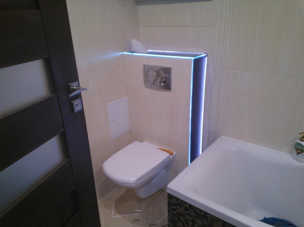 Nowoczesna łazienka z podwieszanym WC i podświetleniem LED w zabudowie stelaża, widoczna wanna z ozdobnym panelem i fragment drzwi.