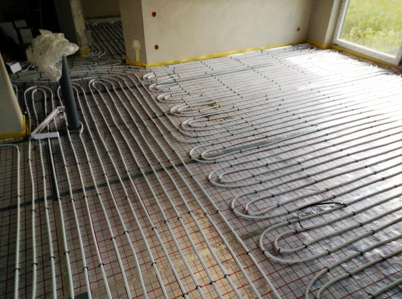 Instalacja ogrzewania podłogowego z białych rurek na czerwonej siatce, zabezpieczona żółtą taśmą na ścianach w Lęborku, widok z góry.